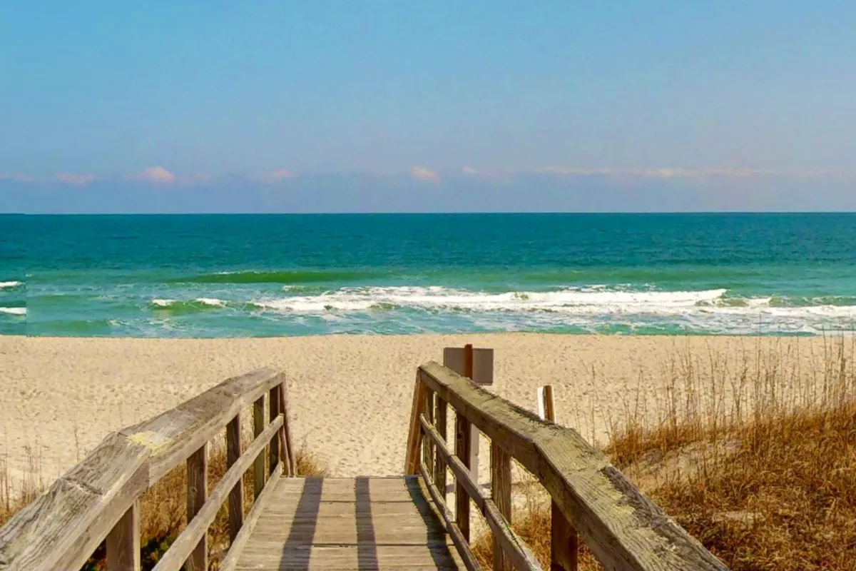 A relaxing scene featuring a wooden boardwalk leading over dune grass to a stretch of sandy beach, with gentle ocean waves in the distance. The setting suggests easy walks to the beach, sandcastle building, and sunbathing in a peaceful, inviting environment.
