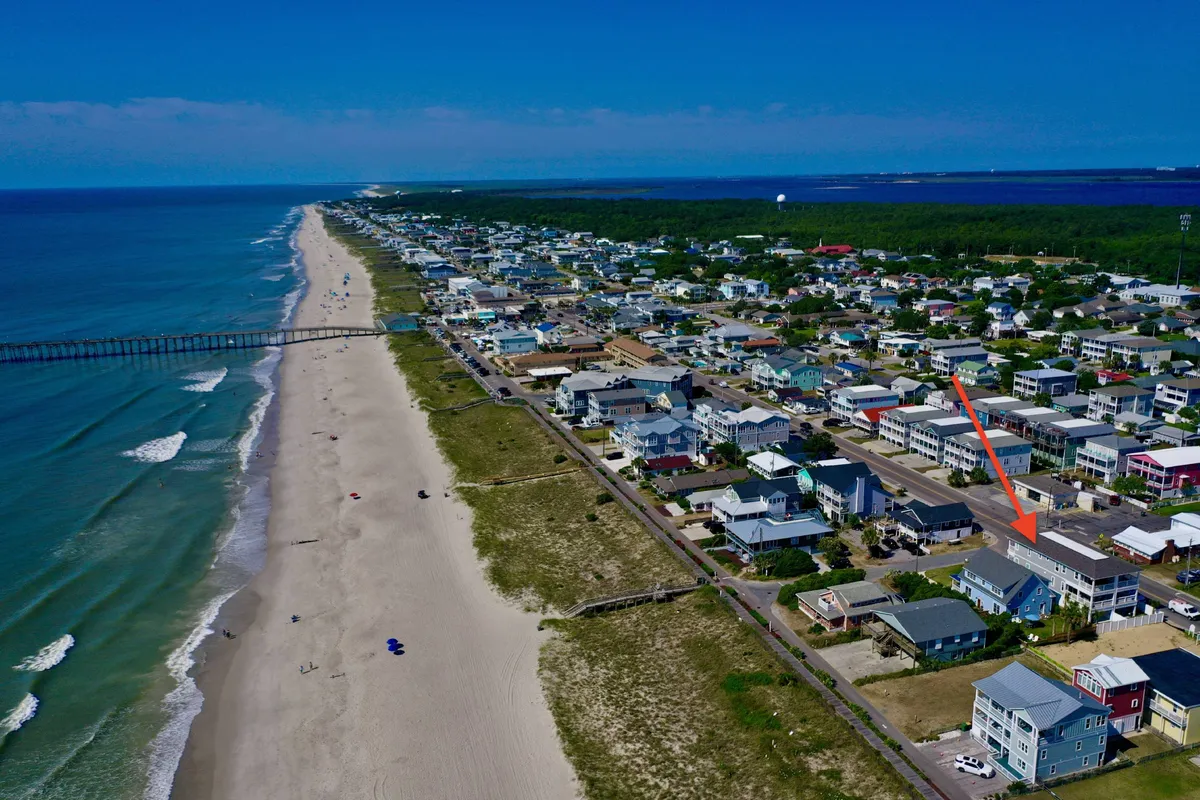 A wide-angle view of the ocean, sandy beach, a pier extending into the water, and nearby houses lining the coast. The scene is lively, with potential for beach activities like sunbathing, fishing, or walking along the shore.