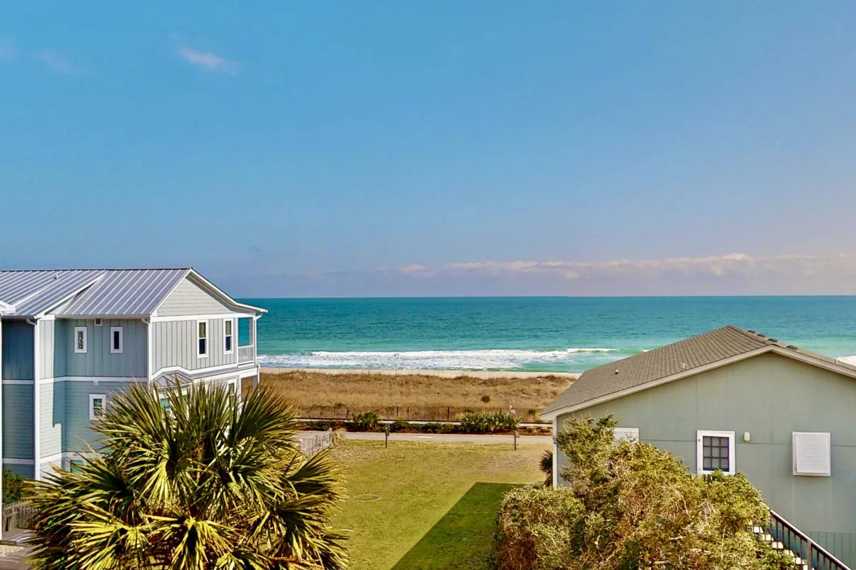 The image showcases a breathtaking ocean view framed by swaying palm trees, with the golden sands of Kure Beach stretching out toward the horizon. In the foreground, you can see the charming townhomes and the inviting beach just steps away, promising easy access to sun, surf, and relaxation. The tranquil setting suggests leisurely beach walks, seaside picnics at sunset, and the gentle sound of waves.