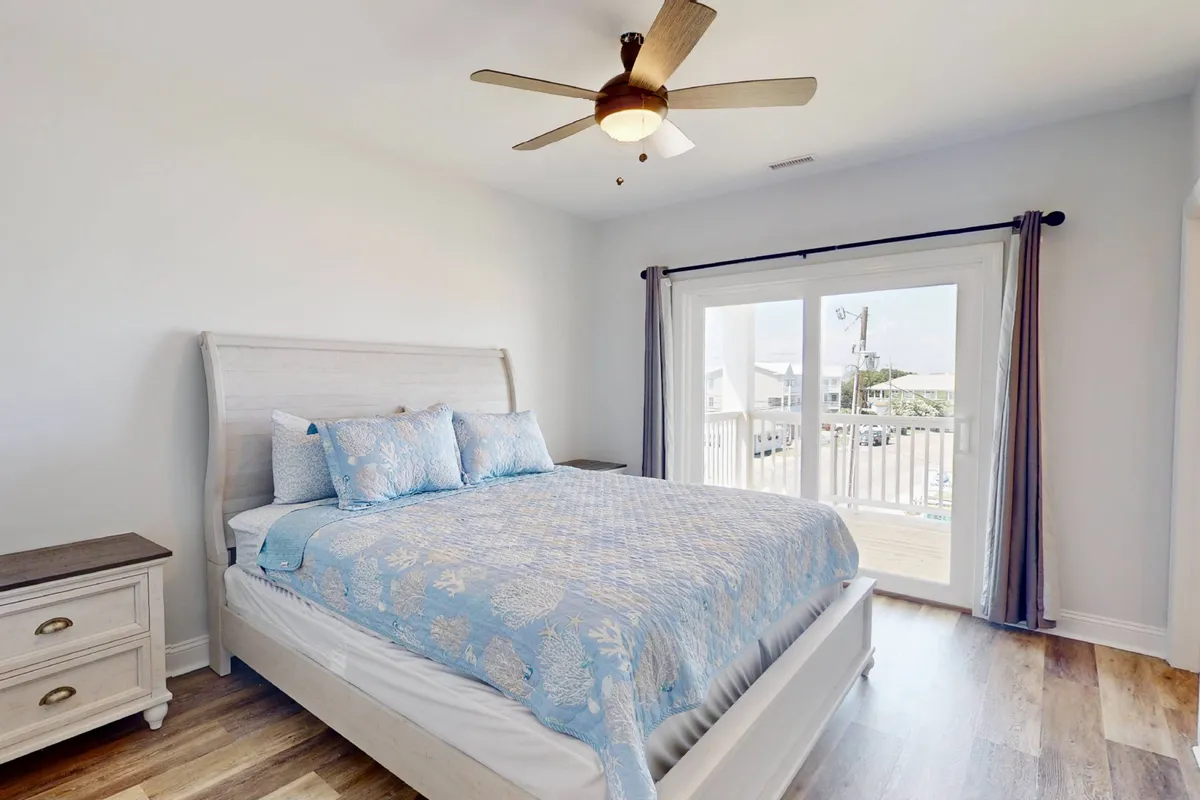 A peaceful bedroom featuring a bed with blue patterned bedding, a large window with a sliding door leading outside, and a nightstand. The room is filled with natural light and has a calming, airy vibe.