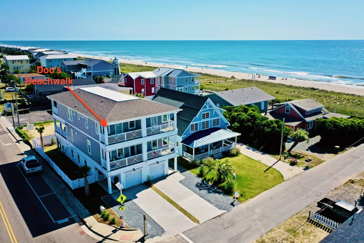 This image provides a broad, cheerful view of the beachfront property, showcasing the proximity to the ocean, a clear beach access path, and several colorful houses lining the shore. The blue ocean is visible, inviting families to walk directly from the property to the sand without crossing busy roads.