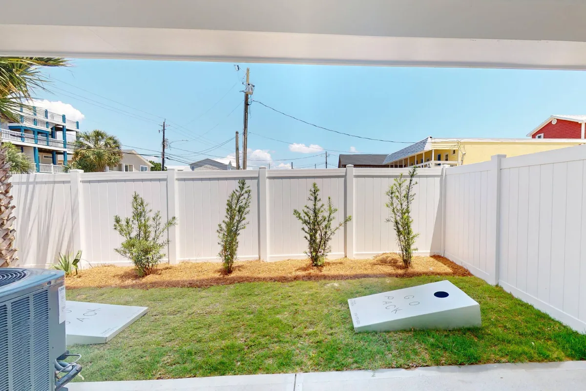 A playful view of the fenced backyard with cornhole boards set up, small shrubs, and sunny open space. This scene conveys opportunities for outdoor fun and family bonding in a safe, private setting.