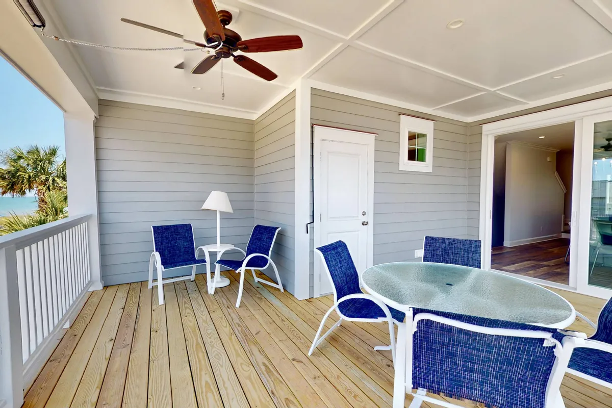This exterior image depicts an outdoor seating area on a covered deck, complete with a glass-topped table, ceiling fan, and a door leading inside. The tranquil and welcoming mood suggests relaxing with a book, enjoying a morning coffee, or hosting small gatherings while taking in the fresh air.
