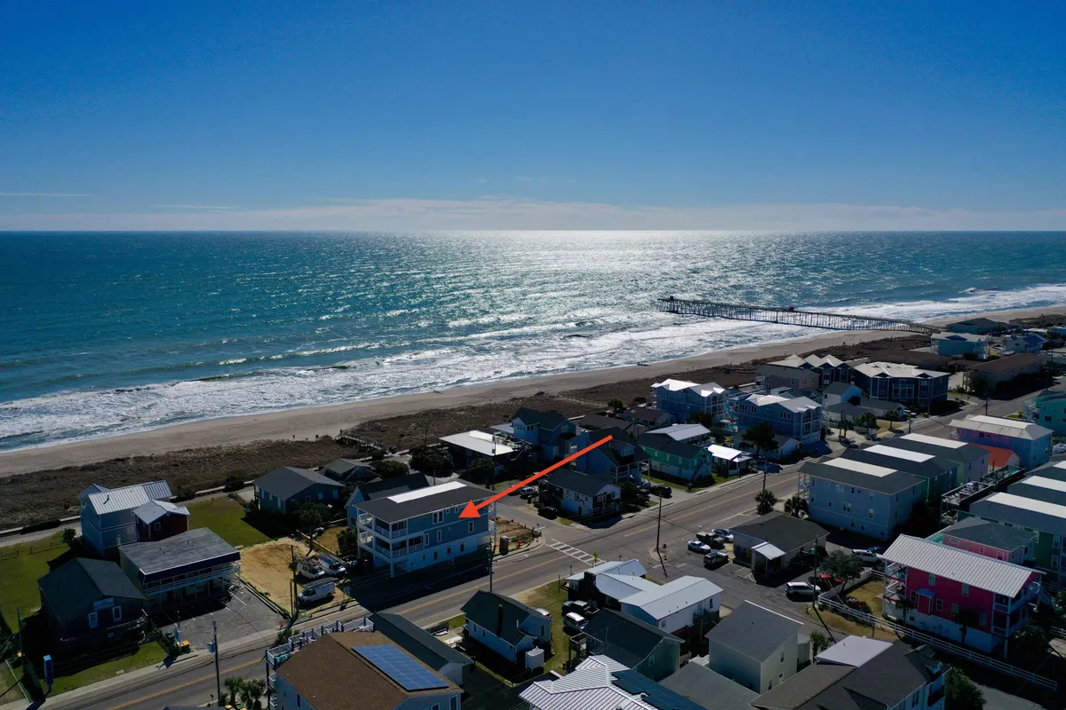 A panoramic view of Kure Beach with the oceanfront, visible pier in the distance, and surrounding neighborhood. The image hints at local attractions like the Kure Beach Pier, opportunities for fishing, ice cream walks, and community activities, all within walking distance.