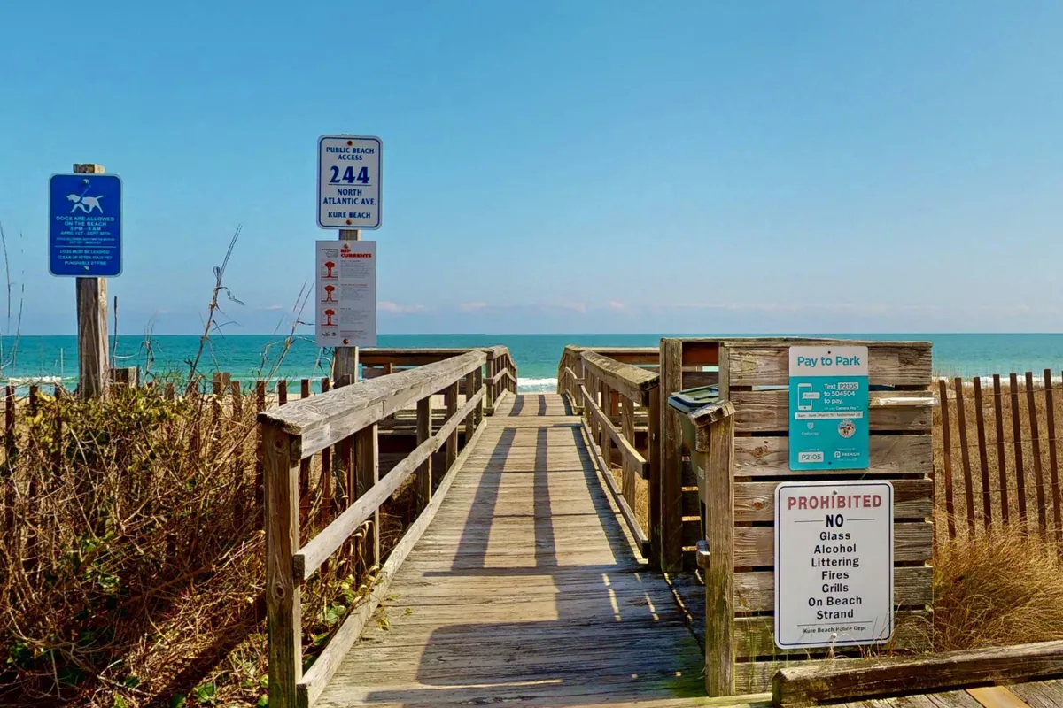 This image captures a wooden boardwalk stretching over sand dunes, leading directly to the ocean. Informational signs dot the scene, and the vast blue ocean is visible in the background. The setting exudes a relaxed and welcoming atmosphere, inviting viewers to imagine themselves heading out for a beach day.