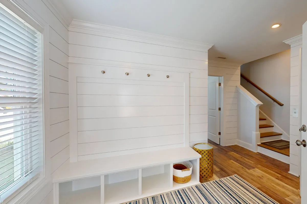 A welcoming entryway featuring a bench with hooks for coats and bags, an area rug, and the base of a staircase. The space is practical for storing shoes and beach gear, while also providing a warm first impression.